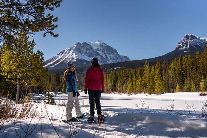 Two friends with snow shoes on a field with snow-covered mountains in the background