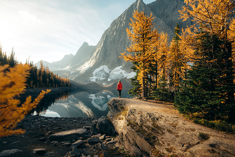Woman at the edge of a rock looking at a lake reflecting the Canadian rockies and Fall colors.